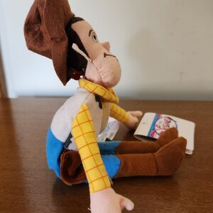 Op de afbeelding: Een pluche speelgoed van Woody uit Toy Story. Hij draagt zijn kenmerkende bruine hoed, gele vest en blauwe jeans. Hij zit met gekruiste benen en uitgestrekte armen.