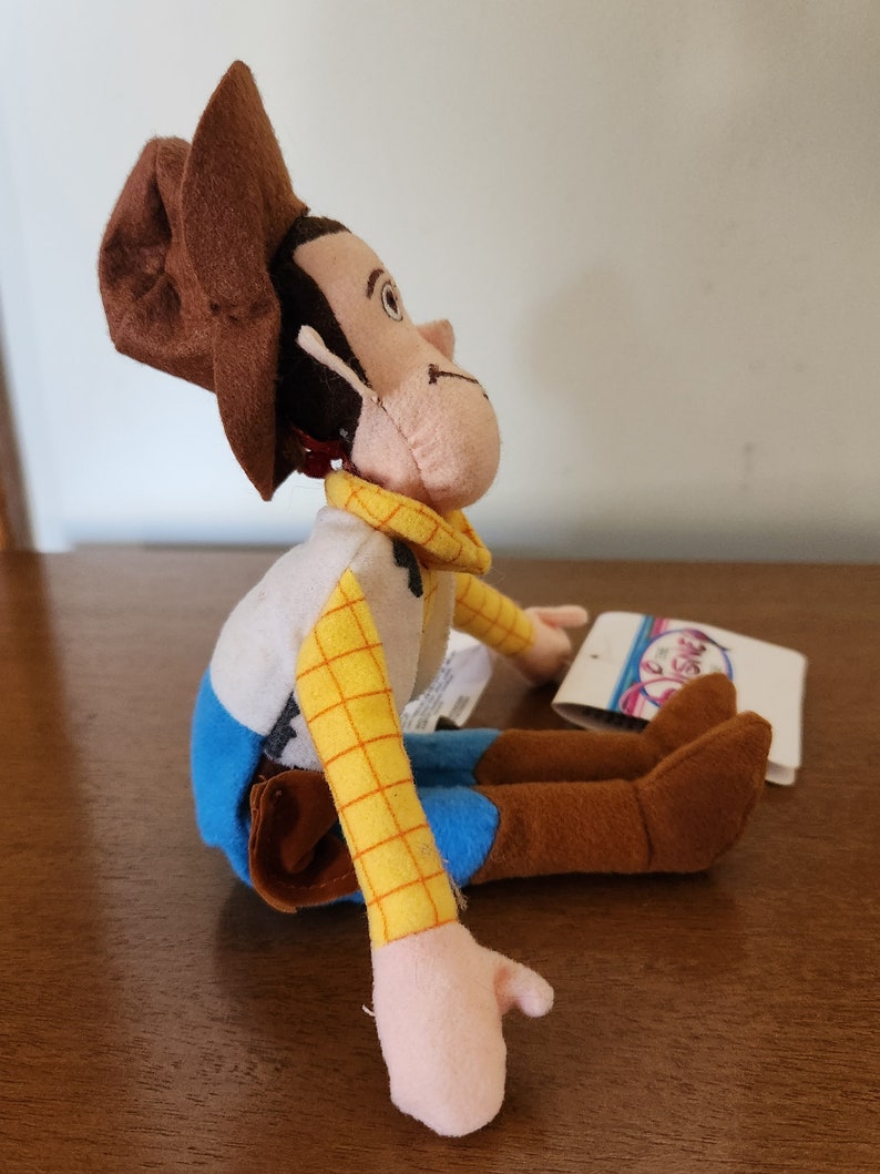 Op de afbeelding: Een pluche speelgoed van Woody uit Toy Story. Hij draagt zijn kenmerkende bruine hoed, gele vest en blauwe jeans. Hij zit met gekruiste benen en uitgestrekte armen.
