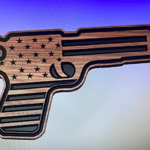 Puede incluir: Un recorte de madera con forma de pistola con un diseño de bandera estadounidense. La pistola es negra y marrón con un patrón de veta de madera.
