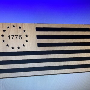 Puede incluir: Un recorte de madera de la bandera estadounidense con 13 rayas negras y un círculo de 13 estrellas que rodean el año 1776.