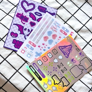 Puede incluir: Tres hojas de pegatinas con varios diseños. La primera hoja tiene un fondo morado con corazones, un murciélago y el texto "my world". La segunda hoja tiene un fondo blanco con corazones rosas y azules, vendas y el texto "Don't fix what ain't broke". La tercera hoja tiene un fondo holográfico con un diseño morado y negro y el texto "Heart-Bit".