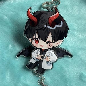 Könnte beinhalten: Ein Schlüsselanhänger mit einem schwarzen und roten Anime-Charakter mit Teufelshörnern und Flügeln. Der Charakter trägt einen weißen Laborkittel und formt mit seinen Händen ein Herz. Der Schlüsselanhänger hat eine silberne Kette und einen silbernen Herzanhänger.