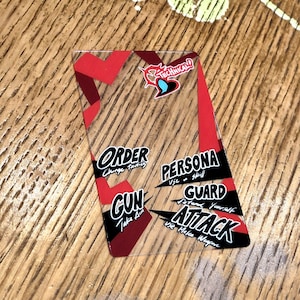 Op de afbeelding: Een doorzichtige plastic kaart met rode en zwarte tekst. De kaart heeft de woorden "ORDER", "PERSONA", "GUARD", "GUN" en "ATTACK" erop. De kaart heeft ook een klein rood hart met een zwarte omtrek en het woord "TECHNICAL!" erop.