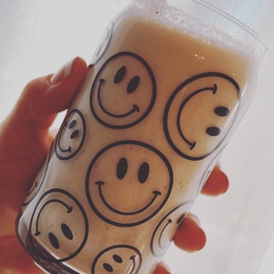 May include: A clear glass can-shaped drinking glass filled with a light-colored beverage. The glass is decorated with black smiley faces. A straw is visible in the drink. The glass is held by a person.