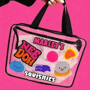 May include: A clear rectangular plastic bag with black trim and two black handles. The bag features colorful stickers including the text MARLEY'S NEE DOH SQUISHIES, a blue round character, a purple cat, a pink circle, a yellow pizza slice, and a gray dumpling.