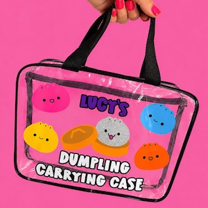 May include: A clear rectangular carrying case with black trim and handles. The front features colorful cartoon dumplings in pink, yellow, blue, and orange, along with a glittery gray dumpling in a steamer. Text reads: LUCY'S DUMPLING CARRYING CASE.