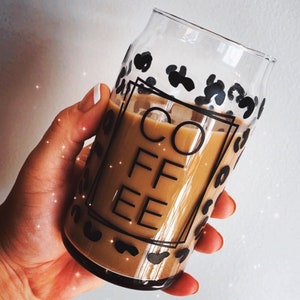 May include: A clear glass coffee cup with a leopard print design and the word "COFFEE" in black block letters. The cup is filled with a light brown beverage, possibly coffee or a latte.