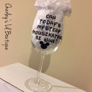 May include: A clear wine glass with black text that reads "Can Today's Mystery Mousekatool Be Wine?" and a black Mickey Mouse head. The glass is filled with white tissue paper.