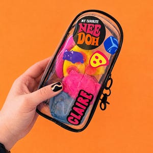 May include: A clear, rectangular case containing colorful Nee Doh stress balls and shapes. The case has a black border and a black clip. The text "MY FAVORITE NEE DOH" and "CLAIRE" are visible on the case.