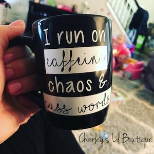 May include: Black ceramic mug with white text that reads "I run on caffeine, chaos & cuss words." The mug has a black handle and a white stripe design. The text is in a handwritten style. The bottom of the mug has the text "Charley's Lil Boutique."