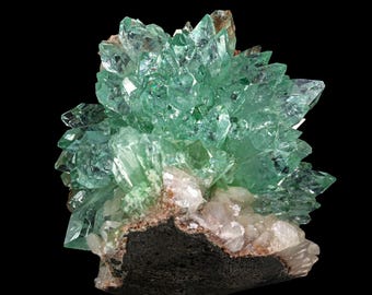 May include: A cluster of green crystals on a dark grey rock. The crystals are sharp and pointed, and they have a translucent appearance.