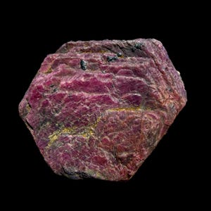 May include: A raw ruby gemstone with a deep red hue, displaying a rough, textured surface. The stone has a hexagonal shape and is set against a black background. The ruby's surface has visible lines and small dark inclusions.