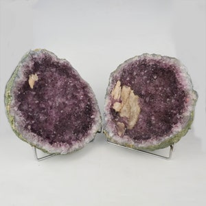May include: Two halves of an amethyst geode, showcasing a deep purple crystal interior. The outer shell is a muted green, and the geode is displayed on a silver metal stand. A light beige mineral deposit is visible inside.