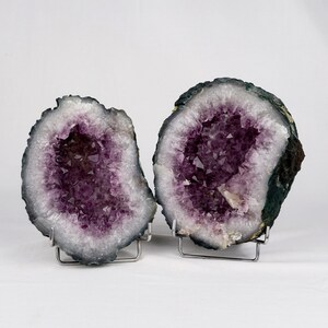 May include: Two amethyst geode halves on metal stands. The geodes are purple and white with a dark green exterior.
