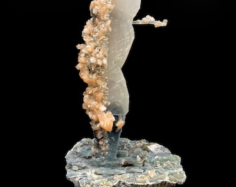 May include: A clear, translucent crystal formation with a cluster of smaller, tan crystals attached to one side. The crystal is standing on a grey, textured base.
