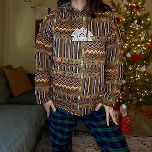 May include: A zip-up hooded jacket with a geometric pattern in shades of brown, orange, and green. The jacket features a white emblem with a mountain design. The person is wearing plaid pajama pants.