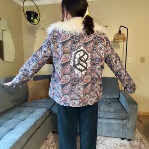 May include: A paisley-patterned jacket with a white faux fur collar. The jacket features a black and white emblem on the back. The pattern includes shades of blue, red, and white. The person is wearing dark blue pants.