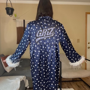 May include: A navy blue robe with a white star pattern and the word "GRIZ" on the back. The robe has long sleeves with white feather trim at the cuffs. The robe is knee-length.