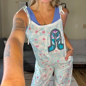 May include: A light blue and white floral jumpsuit with a butterfly graphic on the front. The jumpsuit has thin straps and a lace trim around the neckline.