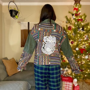 May include: A colorful jacket with patterned sleeves and back panel, featuring a white embroidered design. The jacket has olive green sleeves and collar. The person is wearing blue and green plaid pajama pants.