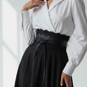 K&ouml;nnte beinhalten: Ein lang&auml;rmeliges Kleid mit einem wei&szlig;en Oberteil mit Kragen in Wickeloptik und einem schwarzen Rock. Die Taille wird durch einen breiten schwarzen G&uuml;rtel mit Spitzenbesatz und einer mittigen Bindung betont, was einen Kontrast zwischen Ober- und Unterteil bildet.
