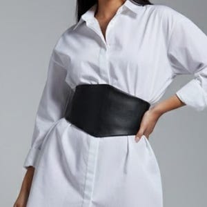 K&ouml;nnte beinhalten: Ein wei&szlig;es Hemdblusenkleid mit Knopfleiste, langen &Auml;rmeln und klassischem Kragen, kombiniert mit einem breiten schwarzen Korsettg&uuml;rtel in der Taille. Das Kleidungsst&uuml;ck wird mit schwarzen Overknee-Stiefeln getragen, was eine klare Silhouette ergibt.