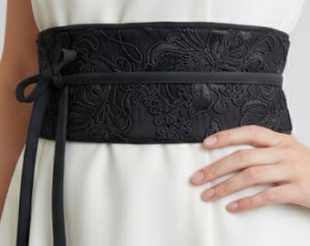 Könnte beinhalten: Ein breiter schwarzer Gürtel im Obi-Stil mit aufwendiger Blumenstickerei und glatter Stoffoberfläche. Der Gürtel ist um ein einfarbiges weißes ärmelloses Kleid gewickelt und wird vorne mit einem dünnen schwarzen Band in zwei langen Streifen gebunden.
