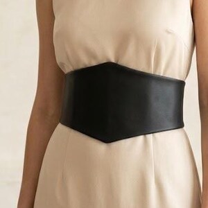 K&ouml;nnte beinhalten: Ein &auml;rmelloses beiges Kleid mit Rundhalsausschnitt und einem breiten, schwarzen Korsettg&uuml;rtel mit spitzer Vorderseite, der die Taille betont. Das Kleid besteht aus einem glatten, einfarbigen Stoff, der gerade von der Taille abf&auml;llt.