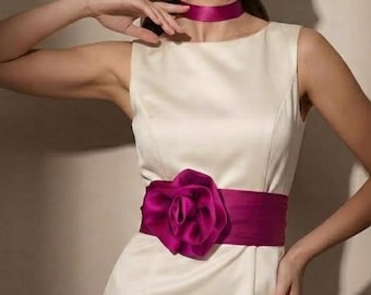 Könnte beinhalten: Ein ärmelloses, cremefarbenes Kleid mit glatter, satinartiger Oberfläche, rundem Ausschnitt und einem breiten, leuchtend magentafarbenen Stoffgürtel in der Taille. Der Gürtel ist mit einer großen, passenden Stoffblume und einem Halsband verziert.
