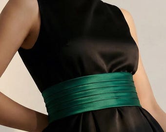 May include: A sleeveless black dress featuring a wide, pleated emerald green satin sash at the waist. The dress has a high, rounded neckline and a full, structured skirt that falls from the waistline, crafted from a smooth, lustrous fabric.