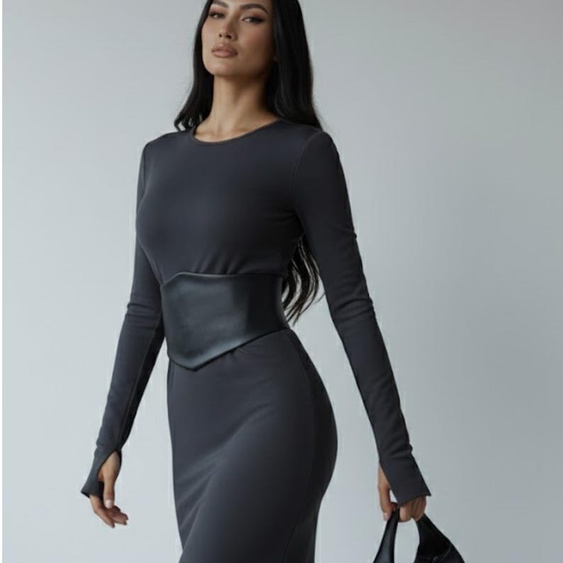 K&ouml;nnte beinhalten: Ein lang&auml;rmeliges, anthrazitfarbenes Midikleid mit Rundhalsausschnitt und Daumenl&ouml;chern an den &Auml;rmelb&uuml;ndchen. Das Kleid wird in der Taille durch einen breiten, strukturierten schwarzen Korsettg&uuml;rtel aus Kunstleder betont.