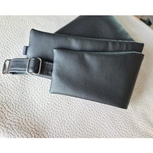 K&ouml;nnte beinhalten: Ein schwarzes Accessoire in Lederoptik mit einer rechteckigen Tasche, die an einem Riemen mit einer silberfarbenen Metallschnalle und einem Schieber befestigt ist. Das Material hat eine glatte, matte Oberfl&auml;che mit sichtbarer Narbung.