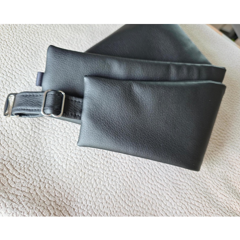 K&ouml;nnte beinhalten: Ein schwarzes Accessoire in Lederoptik mit einer rechteckigen Tasche, die an einem Riemen mit einer silberfarbenen Metallschnalle und einem Schieber befestigt ist. Das Material hat eine glatte, matte Oberfl&auml;che mit sichtbarer Narbung.