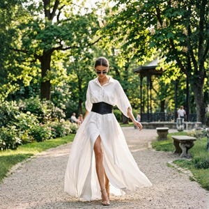 K&ouml;nnte beinhalten: Ein lang&auml;rmeliges wei&szlig;es Hemdkleid mit durchgehender Knopfleiste, einem asymmetrischen Saum mit tiefem Schlitz vorne und einem breiten schwarzen Korsettg&uuml;rtel, der die Taille betont. Das Kleidungsst&uuml;ck wird mit einer dunklen Sonnenbrille kombiniert.