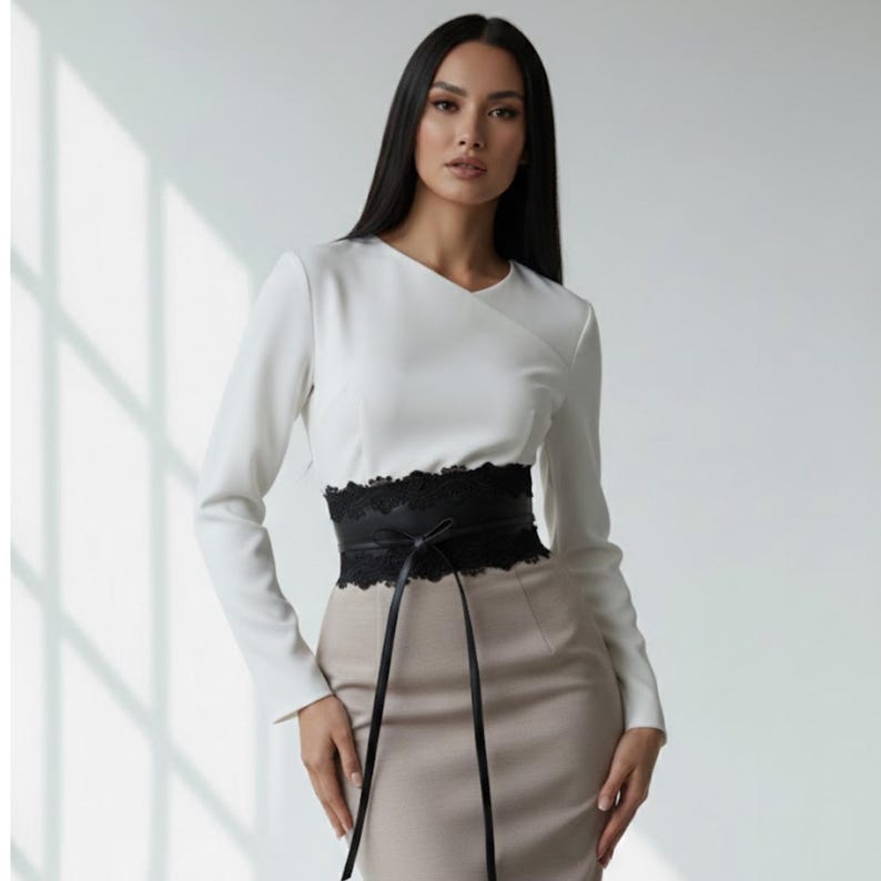 K&ouml;nnte beinhalten: Eine lang&auml;rmelige wei&szlig;e Bluse mit &uuml;berkreuztem Ausschnitt kombiniert mit einem beigefarbenen Rock mit hoher Taille. Ein breiter schwarzer Korsettg&uuml;rtel mit aufwendiger Spitzenborte und d&uuml;nnen Bindeb&auml;ndern wird in der Taille getragen.