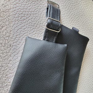 K&ouml;nnte beinhalten: Eine schwarze, texturierte Tasche mit einem passenden verstellbaren Riemen und einer silberfarbenen Metallschnalle. Die Tasche hat eine rechteckige Form mit genarbter Oberfl&auml;che und ist an einem langen, schmalen schwarzen Riemen mit Metallschieber befestigt.