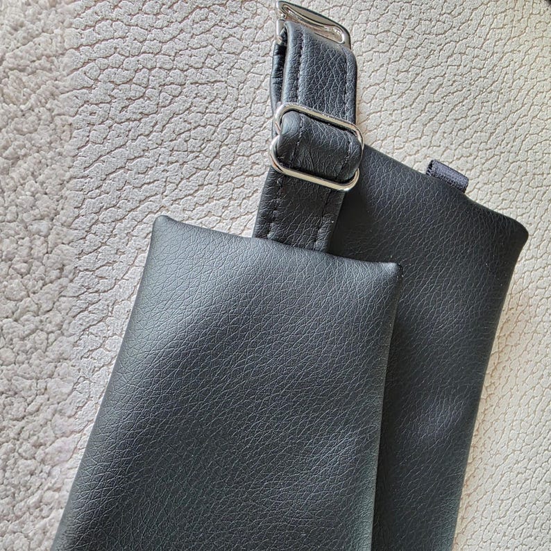 K&ouml;nnte beinhalten: Eine schwarze, texturierte Tasche mit einem passenden verstellbaren Riemen und einer silberfarbenen Metallschnalle. Die Tasche hat eine rechteckige Form mit genarbter Oberfl&auml;che und ist an einem langen, schmalen schwarzen Riemen mit Metallschieber befestigt.
