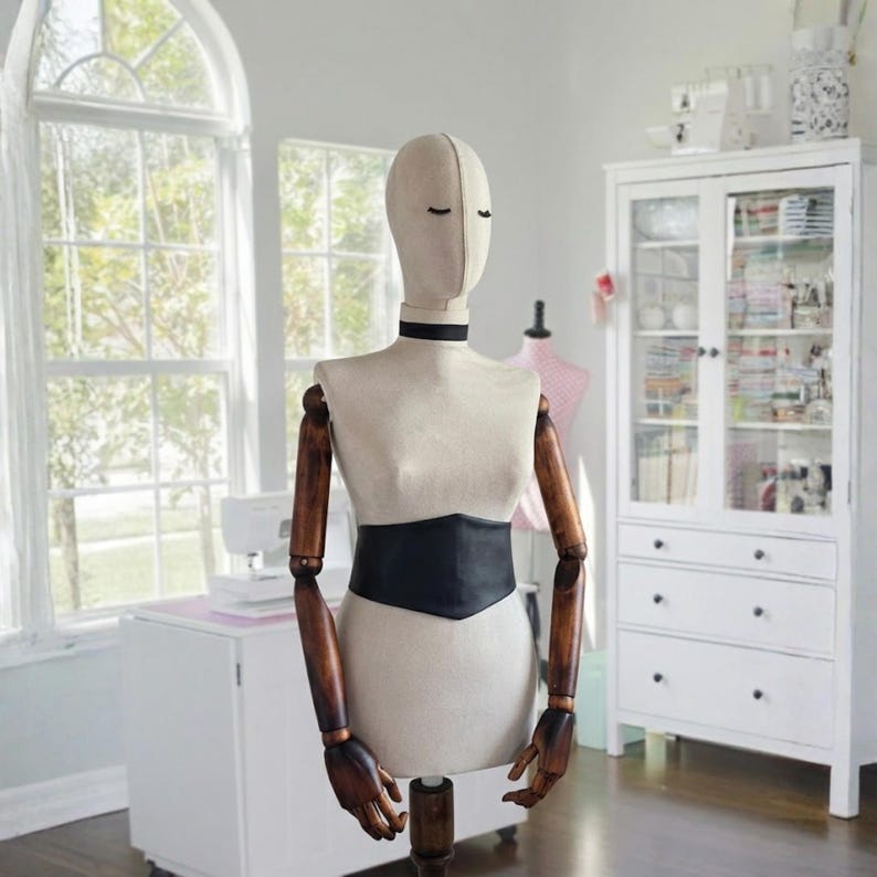 K&ouml;nnte beinhalten: Eine beige Schneiderpuppe aus Stoff mit einem glatten, gesichtslosen ovalen Kopf und einem schwarzen Halsband. Der Torso ist mit einem breiten, schwarzen Tailleng&uuml;rtel versehen. Die Puppe hat bewegliche, dunkelbraune Holzarme und H&auml;nde.