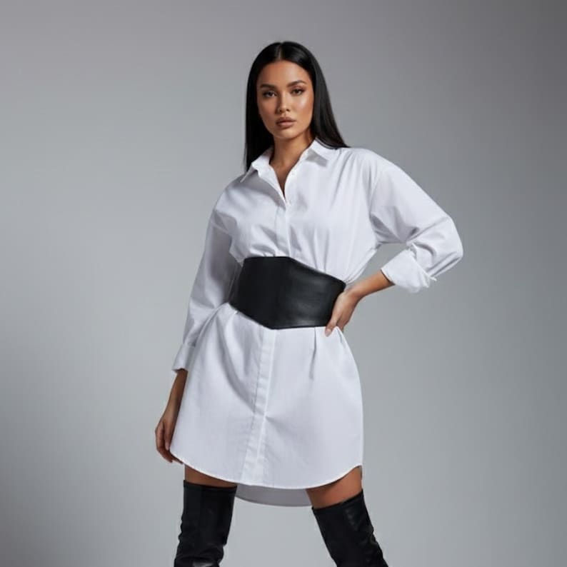 K&ouml;nnte beinhalten: Ein wei&szlig;es Hemdblusenkleid mit Knopfleiste, langen &Auml;rmeln und klassischem Kragen, kombiniert mit einem breiten schwarzen Korsettg&uuml;rtel in der Taille. Das Kleidungsst&uuml;ck wird mit schwarzen Overknee-Stiefeln getragen, was eine klare Silhouette ergibt.