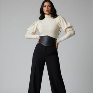 K&ouml;nnte beinhalten: Ein cremefarbenes Langarmshirt mit hohem Stehkragen und volumin&ouml;sen Puff&auml;rmeln, kombiniert mit einer schwarzen, hoch taillierten Hose mit weitem Bein und einem breiten, schwarzen Korsettg&uuml;rtel, der die Taille betont und die Silhouette strukturiert.