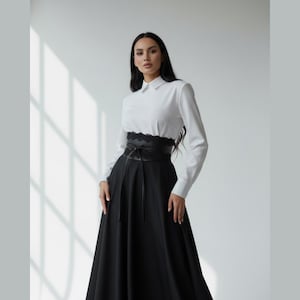 K&ouml;nnte beinhalten: Ein lang&auml;rmeliges wei&szlig;es Hemd mit Kragen kombiniert mit einem bodenlangen schwarzen Rock. Das Outfit verf&uuml;gt &uuml;ber einen breiten schwarzen Tailleng&uuml;rtel mit Spitzenbesatz und Bindeband, was eine strukturierte Silhouette erzeugt.