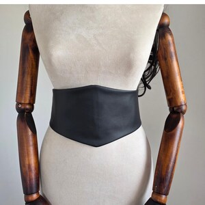 K&ouml;nnte beinhalten: Ein breiter schwarzer G&uuml;rtel im Korsett-Stil mit glatter, matter Oberfl&auml;che und spitz zulaufendem Design an der Vorderseite. Der G&uuml;rtel wird auf einem beigen Schaufensterpuppen-Torso mit beweglichen Holzarmen pr&auml;sentiert und zeigt die einfarbige Optik.