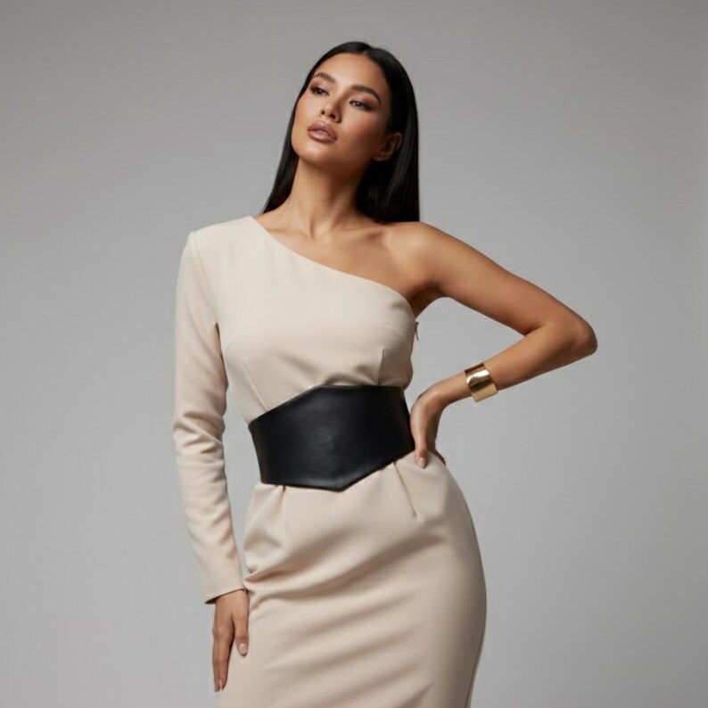 K&ouml;nnte beinhalten: Ein beiges, lang&auml;rmeliges Kleid mit nur einer Schulter und einem breiten, schwarzen Korsett-G&uuml;rtel in Lederoptik. Das Kleid hat eine figurbetonte Passform und wird mit einem gl&auml;nzenden, goldfarbenen Armreif am freien Arm kombiniert.