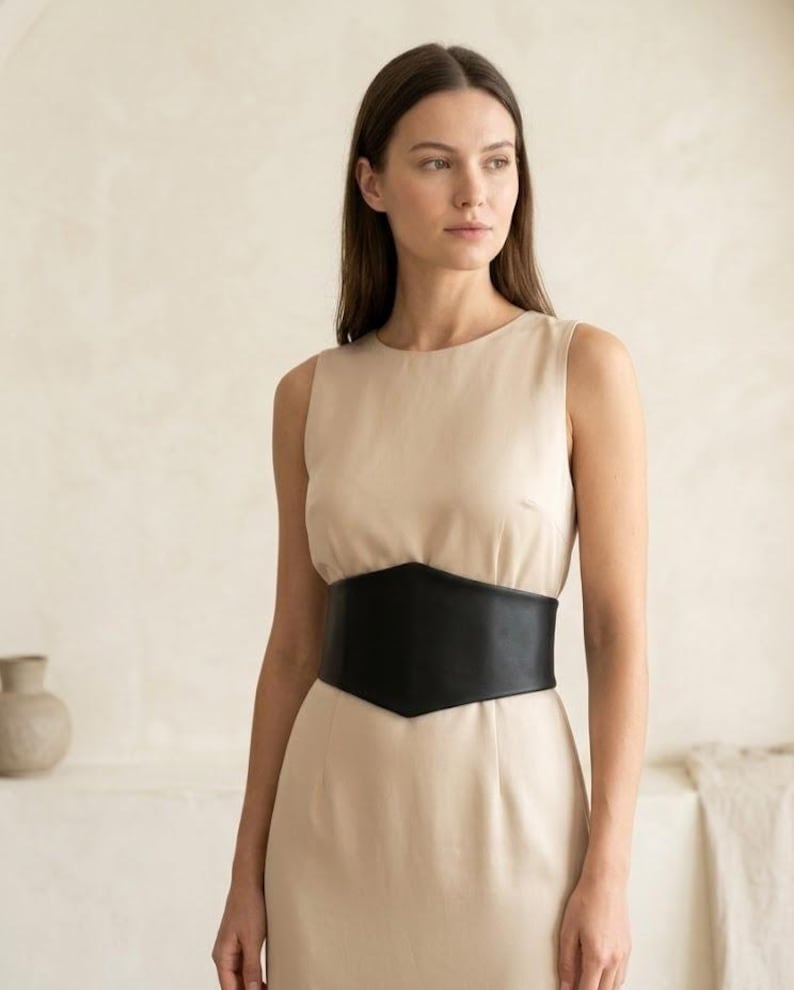K&ouml;nnte beinhalten: Ein &auml;rmelloses beiges Kleid mit Rundhalsausschnitt und einem breiten, schwarzen Korsettg&uuml;rtel mit spitzer Vorderseite, der die Taille betont. Das Kleid besteht aus einem glatten, einfarbigen Stoff, der gerade von der Taille abf&auml;llt.