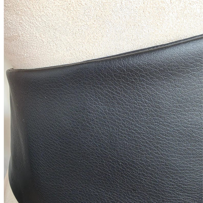 K&ouml;nnte beinhalten: Ein breiter schwarzer G&uuml;rtel mit einer ausgepr&auml;gten genarbten Textur auf der Oberfl&auml;che. Der G&uuml;rtel hat eine glatte, gerade Oberkante und ein gleichm&auml;&szlig;iges mattes Finish, das ein detailliertes Narbenmuster auf dem Material des Accessoires zeigt.
