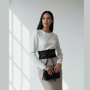 K&ouml;nnte beinhalten: Eine lang&auml;rmelige wei&szlig;e Bluse mit Rundhalsausschnitt kombiniert mit einem beigen Rock mit hoher Taille. Ein breiter schwarzer Korsettg&uuml;rtel aus leder&auml;hnlichem Material mit schwarzer Spitzenborte an den Kanten wird in der Taille getragen.