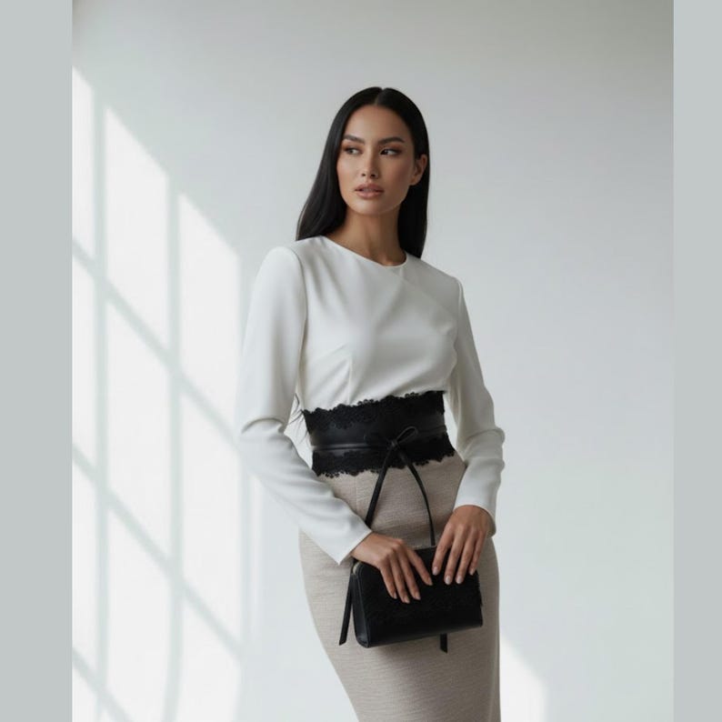K&ouml;nnte beinhalten: Eine lang&auml;rmelige wei&szlig;e Bluse mit Rundhalsausschnitt kombiniert mit einem beigen Rock mit hoher Taille. Ein breiter schwarzer Korsettg&uuml;rtel aus leder&auml;hnlichem Material mit schwarzer Spitzenborte an den Kanten wird in der Taille getragen.