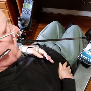 May include: A person in a chair with a black shirt and gray pants is using a long arm to hold a black smartphone. The smartphone is displaying a screen with a blue background and white icons. The person is wearing a black headset with a microphone.