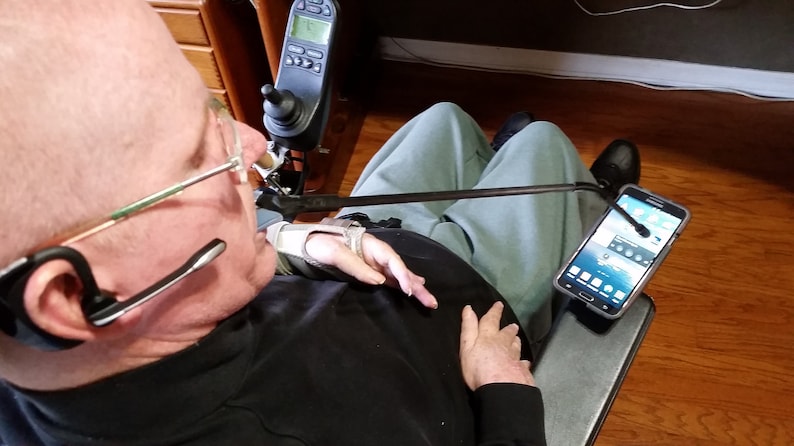 May include: A person in a chair with a black shirt and gray pants is using a long arm to hold a black smartphone. The smartphone is displaying a screen with a blue background and white icons. The person is wearing a black headset with a microphone.