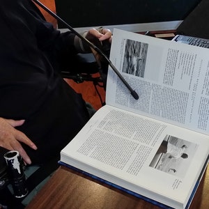May include: A person in a wheelchair is reading a large, open book with a black light attached to a flexible arm. The book has a black and white photo of a bird on the water on the left page and a black and white photo of a person on the right page.